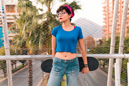 chica patinadora posando con su skate, con maquillaje, lentes y estilo 80s 90s en la ciudad.