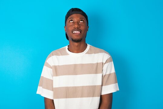 Handsome man wearing striped t-shirt and cap over blue background showing grimace face crossing eyes and showing tongue. Being funny and crazy