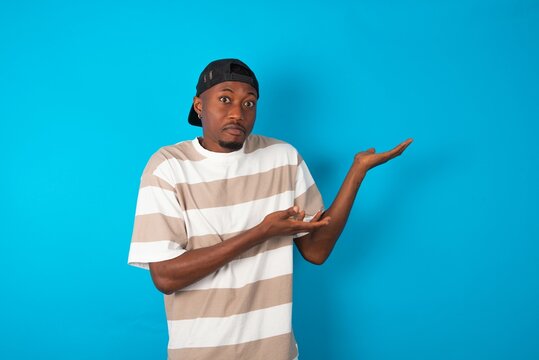 Handsome man wearing striped t-shirt and cap over blue background pointing aside with both hands showing something strange and saying: I don't know what is this. Advertisement concept.