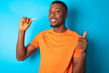 Handsome man wearing  orange T-shirt over blue background holding an invisible braces aligner and...