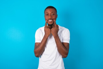 Happy Handsome man wearing white T-shirt over blue background with toothy smile, keeps index fingers near mouth, fingers pointing and forcing cheerful smile