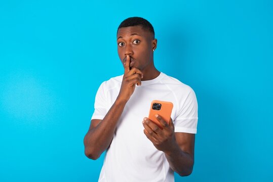 Handsome man wearing white T-shirt over blue background holding modern gadget ask not tell secrets