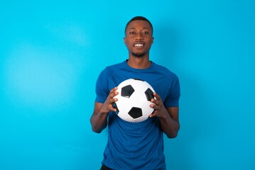 Young man wearing blue T-shirt holding a ball over blue background with nice beaming smile pleased...