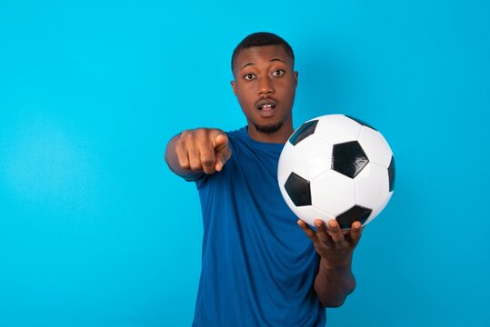 Excited positive Young man wearing sport T-shirt holding a ball over blue background points index finger directly at you, sees something very funny. Wow, amazing - Powered by Adobe