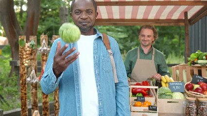Zooming out on two multiethnic people standing at market. Joyful male seller giving cabbage to customer while happy man throwing up natural food and smiling at camera. Recommending foods stall.