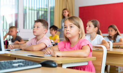 Portrait of focused cute small schoolgirl writing exercises in workbook during lesson in classroom ..