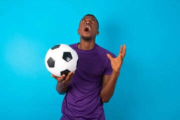 Man wearing purple T-shirt holding a ball over blue background crying and screaming. Human...