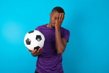 Frustrated Man wearing purple T-shirt holding a ball over blue background holding hand on forehead being depressed regretting what he did having headache, looking stressful.