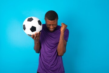 Portrait of Man wearing purple T-shirt holding a ball over blue background being overwhelmed,...