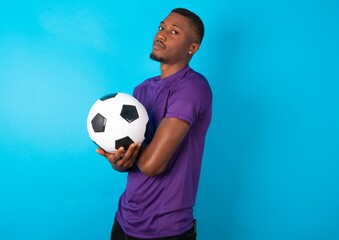 Image of cheerful Man wearing purple T-shirt holding a ball over blue background with arms crossed. Looking and smiling at the camera. Confidence concept.