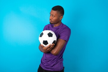 Displeased Man wearing purple T-shirt holding a ball over blue background with bad attitude, arms crossed looking sideways. Negative human emotion facial expression feelings.