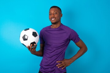 Studio shot of cheerful Man wearing purple T-shirt holding a ball over blue background keeps hand on hip, smiles broadly.