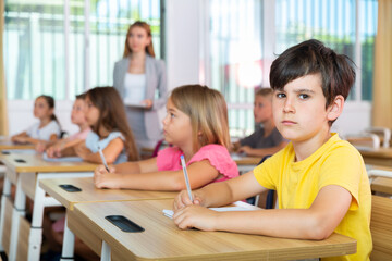 Schoolkids sitting at desks during lesson. Young boy looking in camera