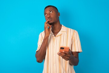 Handsome man wearing fashion shirt over blue background thinks deeply about something, uses modern...
