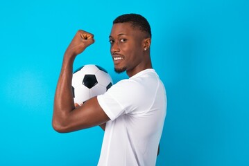 Young man wearing white T-shirt holding a ball over blue background ,  showing muscles after workout. Health and strength concept.