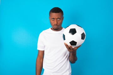 Young man wearing white T-shirt holding a ball over blue background frowning his eyebrows being displeased with something.
