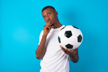 Portrait of thoughtful Young man wearing white T-shirt holding a ball over blue keeps hand under chin, looks away trying to remember something or listens something with interest. Youth concept.