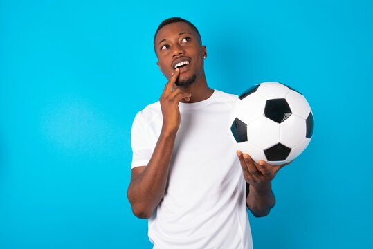 Young man wearing white T-shirt holding a ball over blue background with thoughtful expression, looks to the camera, keeps hand near face, bitting a finger thinks about something pleasant.