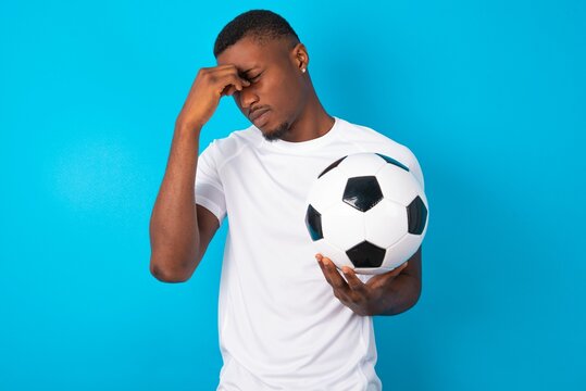 Very upset, Young man wearing white T-shirt holding a ball over blue background touching nose between closed eyes, wants to cry, having stressful relationship or having troubles with work