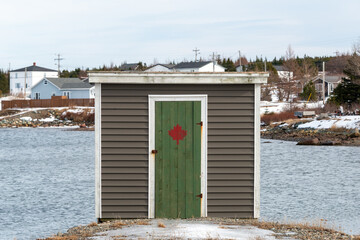 A brown colored small storage shed with a green wooden door. There's a painted red maple leaf on...