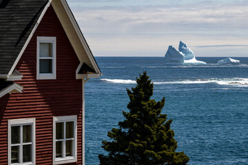 A vibrant red colored two storey cottage style house with glass windows overlooking the blue ocean. In the distance, there's a large white glacier iceberg. The evening sky is pale blue and pink color.