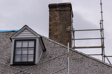 A vintage double hung window and dormer on a beige colored exterior cedar roof of an old building. The windows are black with white trim. There's construction scaffolding in front of the building.