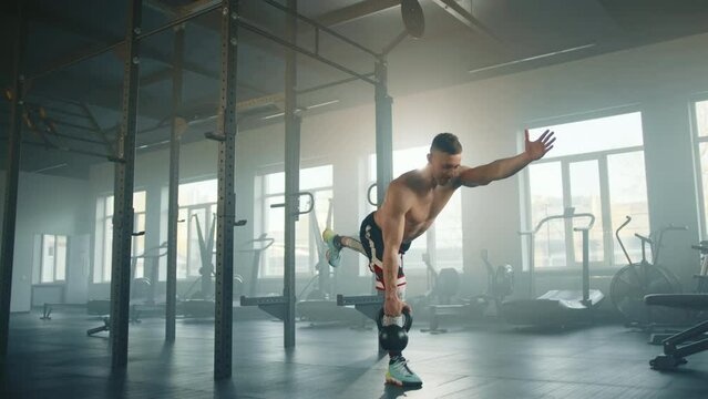 Athletic young man pulls heavy kettlebell up with one hand doing single leg stand in fully-equipped gym. Sportsman holds balance with outstretched arm. High quality 4k footage