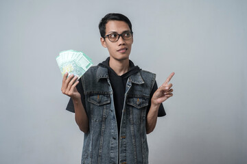 Asian man wearing sleeveless blue jean jacket, happy pose holding rupiah cash. Isolated gray...