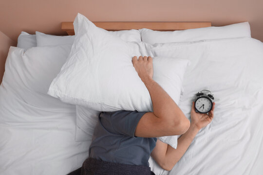 Man covering his face with pillow and holding alarm clock in bed, above view