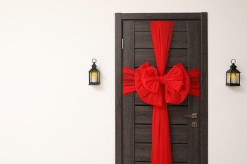 Wooden door with beautiful red bow and lanterns hanging on wall, space for text. Christmas decoration
