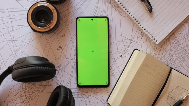 Mobile phone with a green screen and motion capture dots, top view, lies on an abstract painting next to a photo lens, a notebook and headphones. Smartphone Application concept for artists, designers
