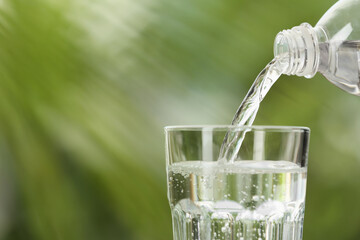 Pouring water from bottle into glass on blurred green background, closeup. Space for text