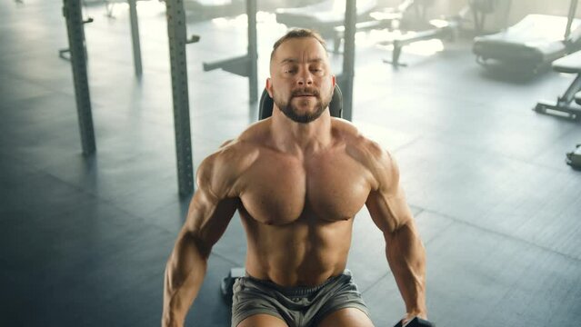 Middle shot of man with naked muscular torso performing lateral arm raise sitting in gym. Man wears boxer briefs. Treadmills, air bikes in the background. High quality 4k footage