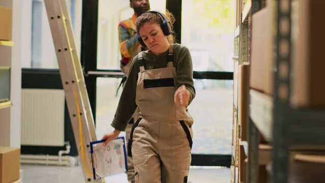 Depot manager having fun in warehouse, reviewing inventory list and listening to music while she works in storage room. Young adult checking retail order shipment, supply chain. Handheld shot.