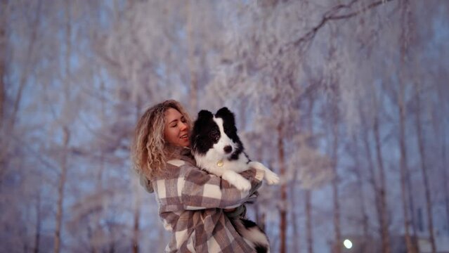 Girl holds a border collie puppy in her arms and kiss in a frosty winter forest covered with frost and snow, at a pink sunset