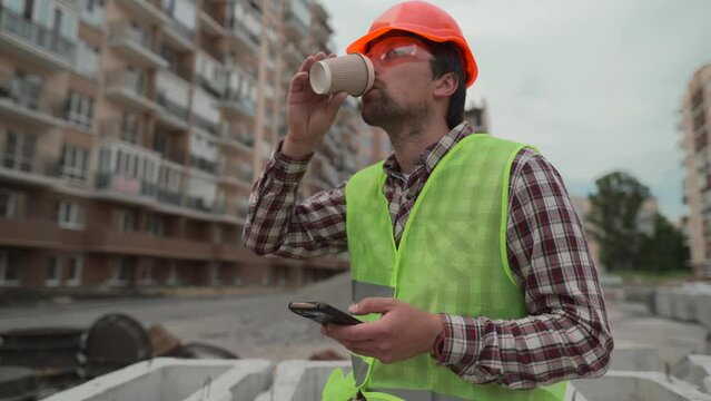 Construction worker resting on building site drinking coffee to go and using smartphone while sitting on jobsite. Architect chatting and using smartphone during coffee break at work project location. 