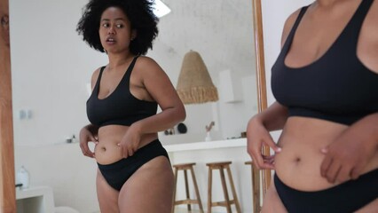 Black woman in front of mirror is upset about her overweight, obesity, unhealthy body. Low self-esteem, critical attitude to own body, internal fat-shaming. Impossibility of accepting one's own body.