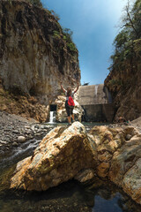 vertical shot of a female hiker with a backpack posing with her arms up in front of a dam in the riverbed on a hot sunny day in the province of Puntarenas in Costa Rica