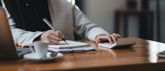 businesswoman or accountant taking note and working on calculator and laptop computer to calculate business data during make note at notepad, accountancy document at office.