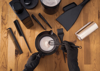 Womens hands in black gloves hold a bowl of paint and prepare tools for hair dyeing. Accessories on a wooden table, top view. Coloring and bleaching of hair at home or in a hair salon.