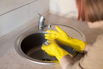 A young girl in yellow rubber gloves washes a glass glass under running tap water in the kitchen. The plot is about housework, cleanliness and daily activities.