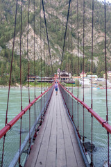 Suspension red bridge across a wide river in the mountains, on which people walk. Safe crossing of the bridge from one bank to another.
