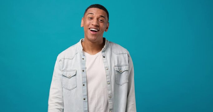 Young african american man waves hand palm in hello gesture welcomes someone expresses positive emotion standing over blue studio background