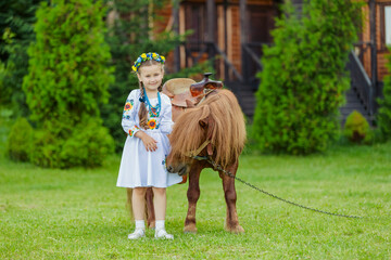 Obraz premium young girl in Ukrainian national dress strokes a pony that grazes on the lawn