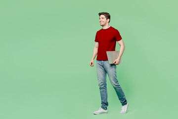 Fototapeta na wymiar Full body side view happy smiling young IT man he wear red t-shirt casual clothes hold closed laptop pc computer go isolated on plain pastel light green background studio portrait. Lifestyle concept.