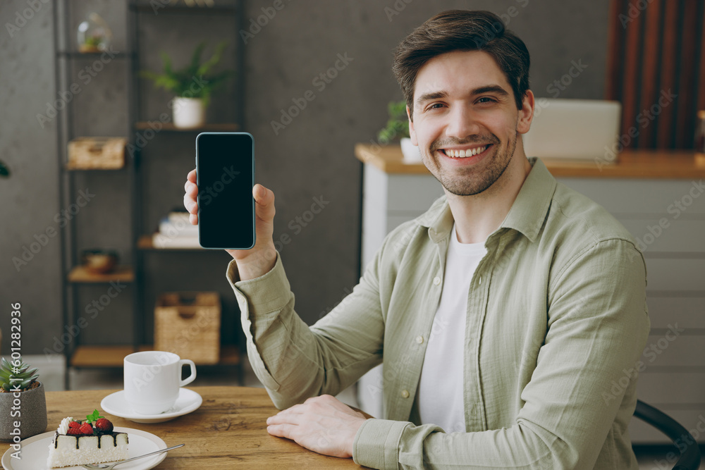 Wall mural Young caucasian man wear casual clothes sits alone at table in coffee shop cafe restaurant indoors work hold use mobile cell phone with blank screen area look aside rest relax during free time indoor.