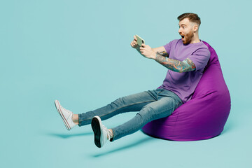 Full body gambling young man in purple t-shirt sit in bag chair use play racing app on mobile cell phone hold gadget smartphone for pc video games isolated on plain pastel light blue cyan background © ViDi Studio