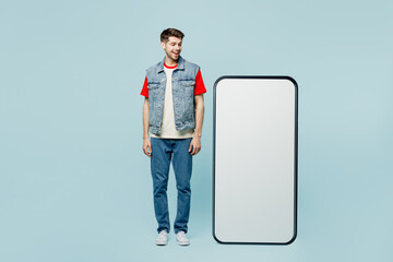 Full body amazed fun young man wear denim vest red t-shirt casual clothes looking at big huge blank screen mobile cell phone smartphone with area isolated on plain pastel light blue cyan background.