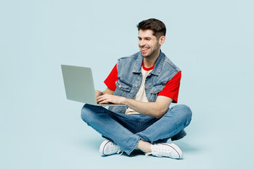 Fototapeta Full body smiling fun young happy IT man wears denim vest red t-shirt casual clothes sit hold use work on laptop pc computer chatting online isolated on plain pastel light blue cyan background studio. obraz