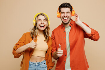 Young fun couple two friends family man woman wear casual clothes headphones listen music show thumb up looking camera together isolated on pastel plain light beige color background studio portrait.